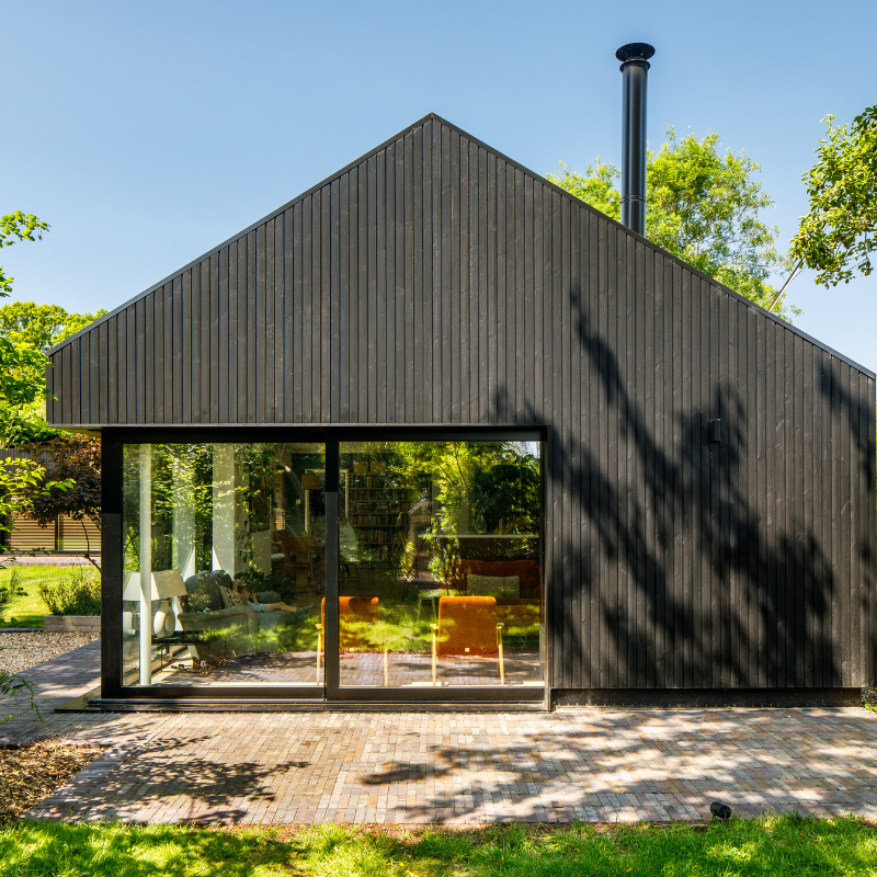 A modern house with a minimalist design. The exterior is finished in Millworks vertical Charcoal Black Thermowood timber cladding. The surrounding greenery contrast with the dark wood, creating a harmonious balance between the building and nature.